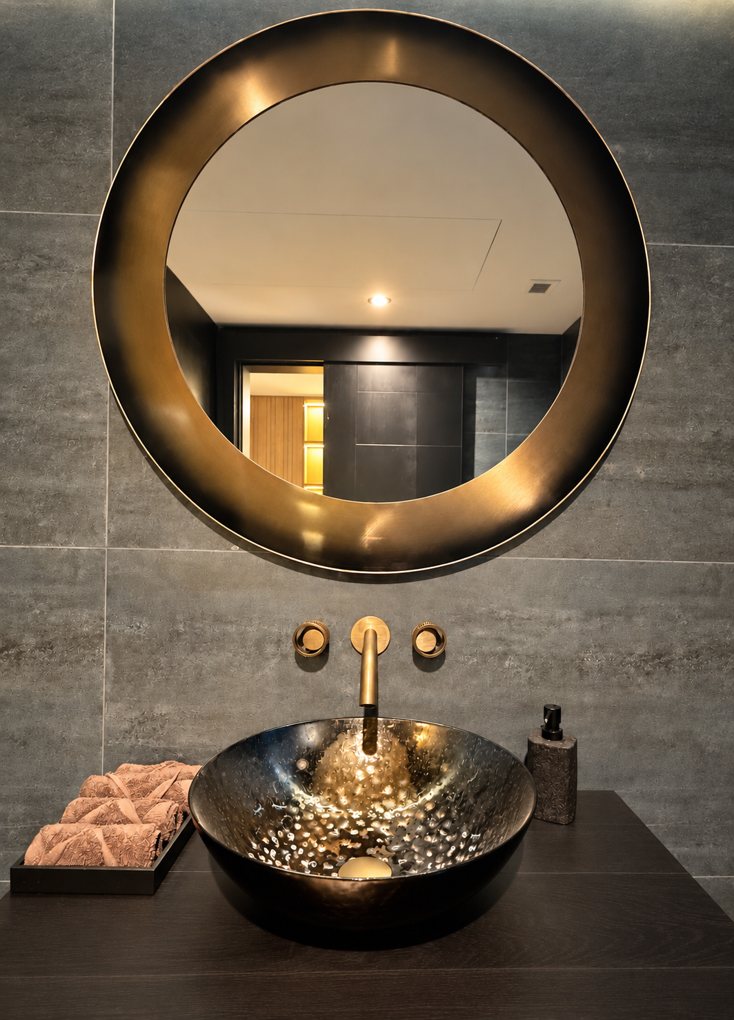 Dark powder room with round illuminated mirror, dark stone wall, brass fittings and black vessel basin