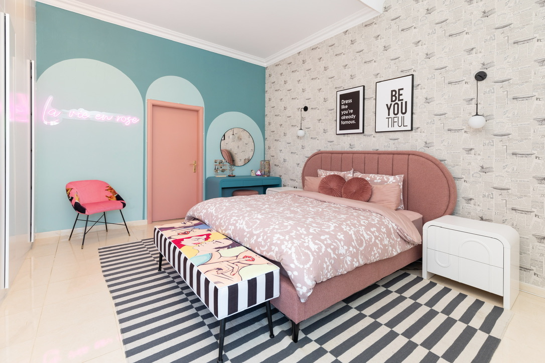 Full bedroom interior with pink bed, turquoise arch accent wall, pink door, turquoise vanity, pink chair, and striped rug