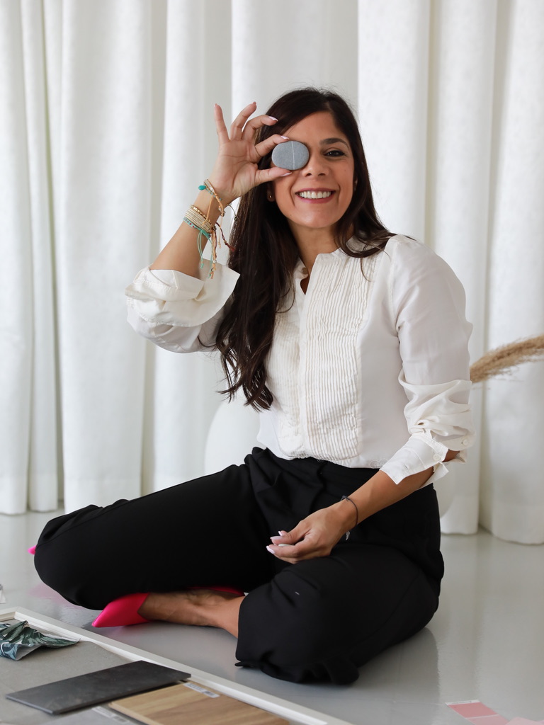 A woman seated cross-legged, smiling, holding a small stone near her eye, wearing an ivory blouse and black trousers, with interior-design material samples nearby