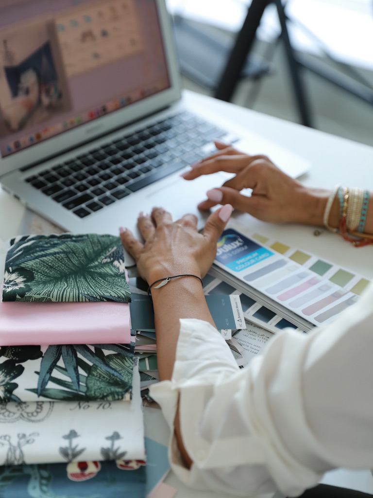 High-angle view of hands resting on paint color swatches and fabric samples beside an open laptop on a white desk.