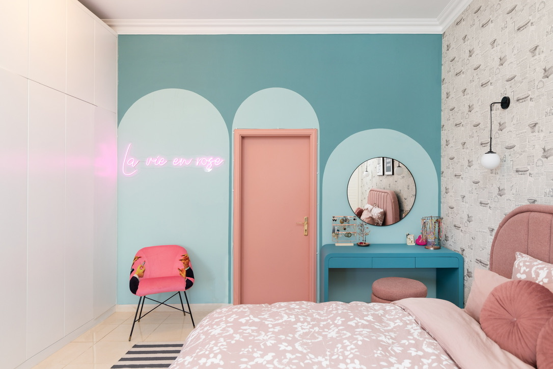 A bedroom featuring a turquoise feature wall with three elegant arches: the left arch frames a pink chair with a glowing pink neon sign reading "la vie en rose" above it, the center arch frames a pink door, and the right arch frames a round mirror above a turquoise vanity. White built-in wardrobes occupy the left wall, patterned wallpaper decorates the right, and a bed with a pink floral duvet is visible in the foreground.