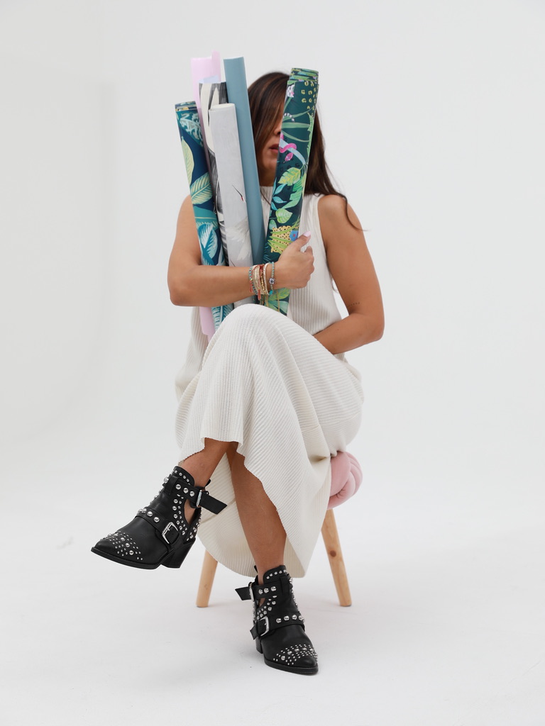 A woman seated on a low stool, leaning forward, holding a bundle of colourful wallpaper rolls against her chest, with crossed legs, boots, and the stool visible below.
