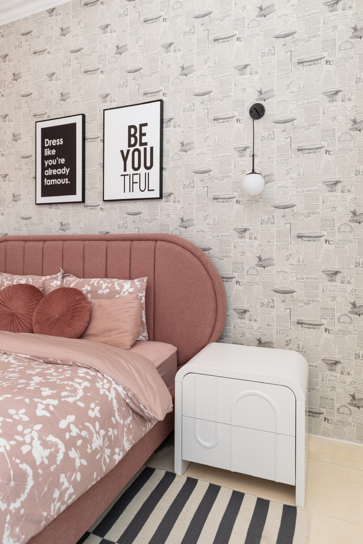 Pink upholstered bed and headboard detail flanked by patterned wallpaper, framed artwork, and white sculptural bedside table