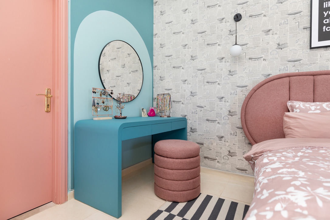 Turquoise vanity dressing area with circular mirror, pink door, pink stool, and view into the bedroom
